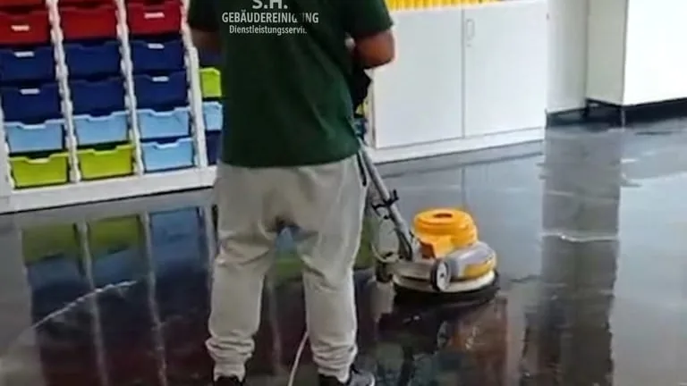 Mitarbeiter im Arbeitsshirt mit S.H.-Logo führt eine Einscheibenmaschine über einen nassen dunklen Boden; im Hintergrund Regale mit farbigen Materialboxen
